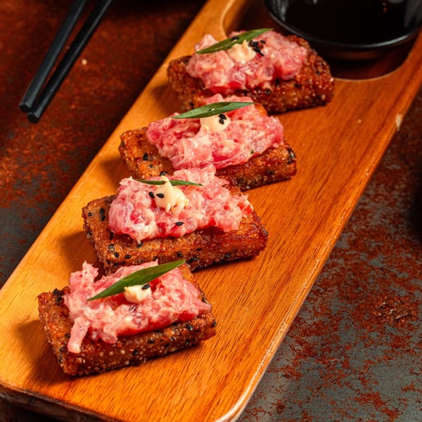 Four SUSHARK sushi bites with pink chopped fish, sauce, and green garnish on a wooden board.